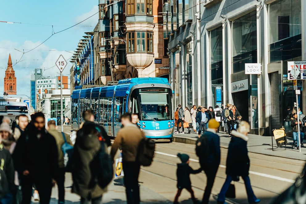 Blue tram on a busy urban street with pedestrians, shops including 'New Look', and a speed limit sign of 15. A red brick building with a tower is in the background